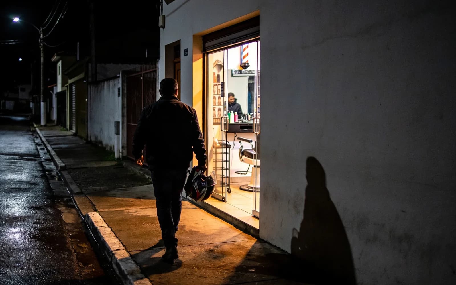 Homem com capacete no braço chegando à porta iluminada de uma barbearia à noite. Imagem ilustrativa.
