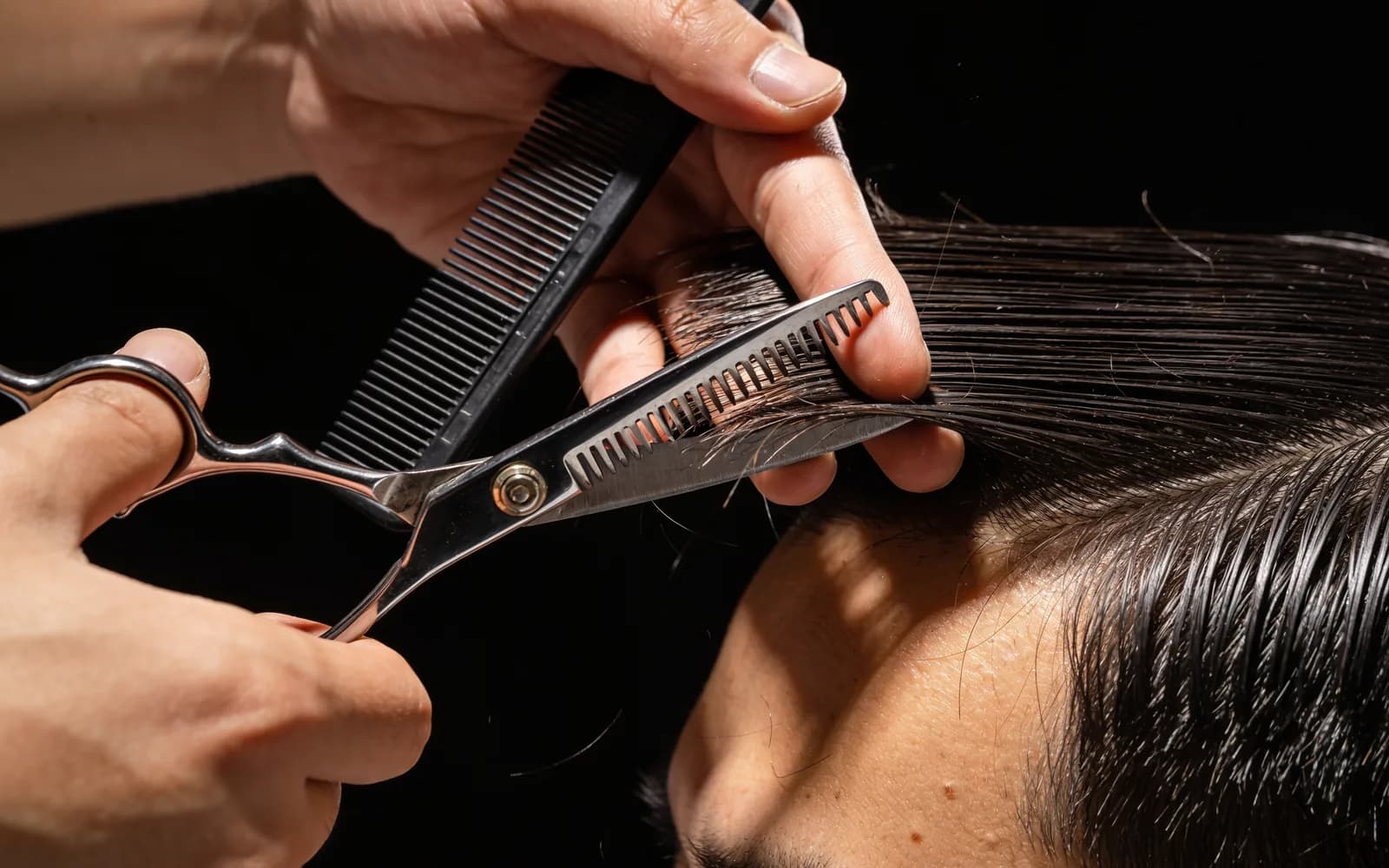 Mãos do barbeiro cortando cabelo masculino com tesoura e pente, em close, sob luz lateral dura. Imagem ilustrativa.